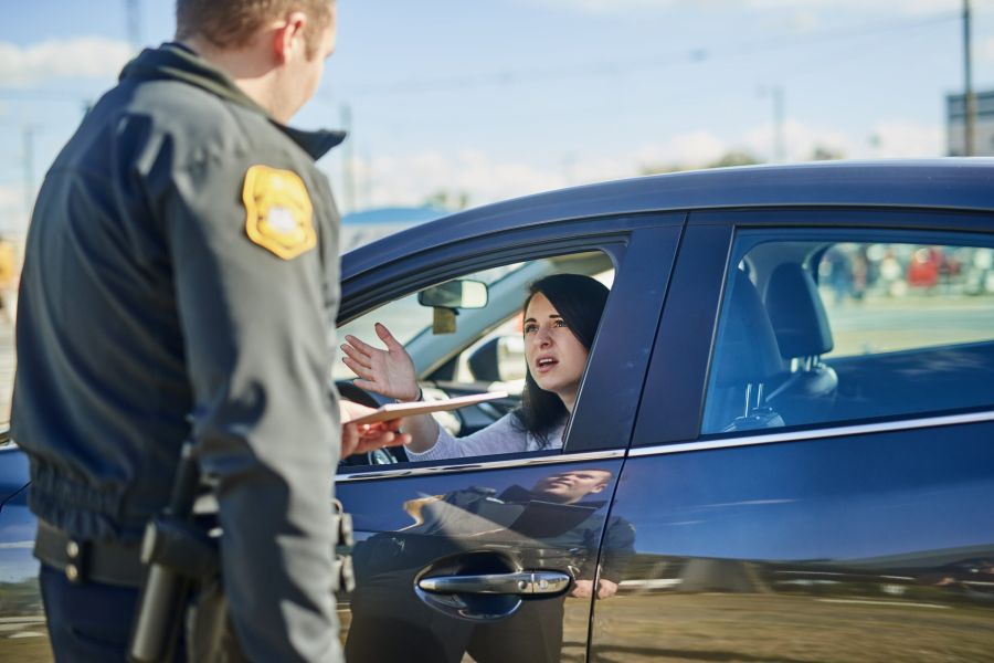 A police officer standing beside a car, speaking to a woman in the driver's seat who appears to be explaining something.