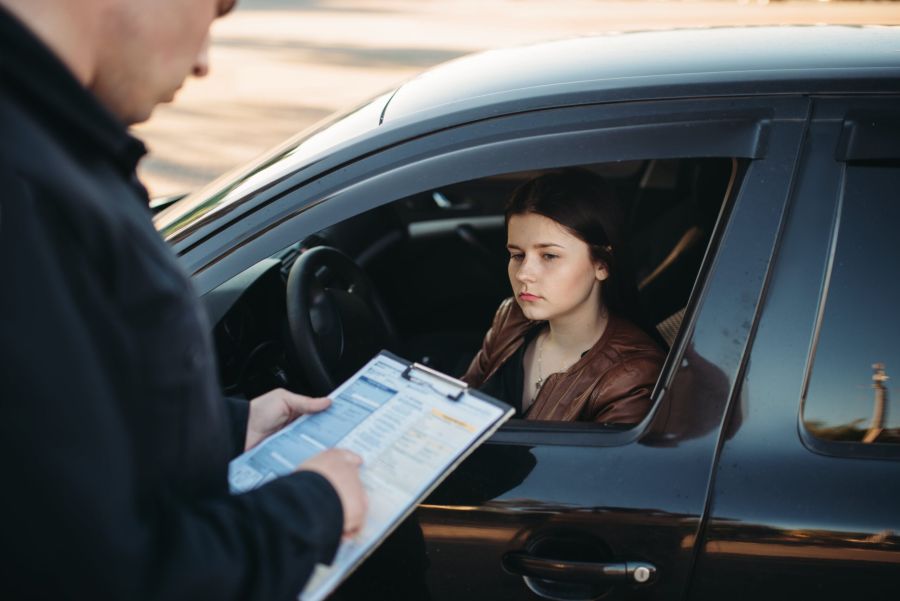 Female driver sitting in a car looking serious as a police officer writes a ticket on a clipboard.