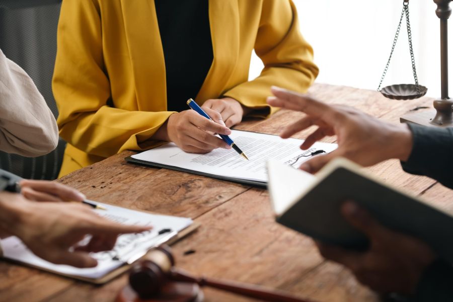 A lawyer in a yellow blazer reviews a document at a wooden desk, with a gavel and scales of justice in the background.