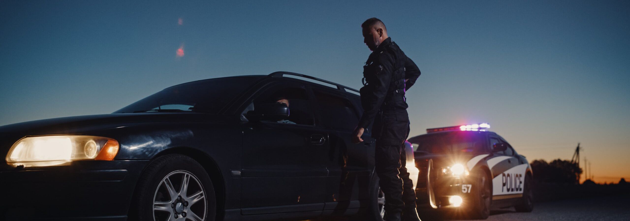 A police officer standing next to a pulled-over car at dusk, with a police vehicle flashing its lights in the background.