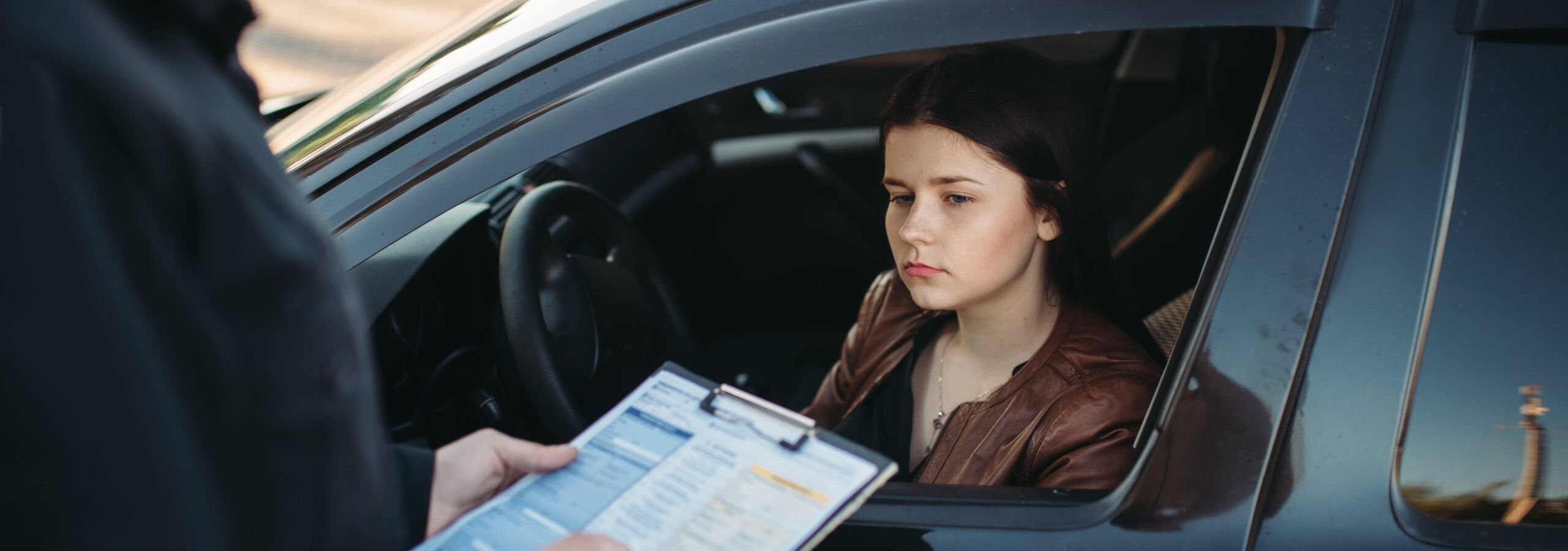 Female driver sitting in a car looking serious as a police officer writes a ticket on a clipboard.